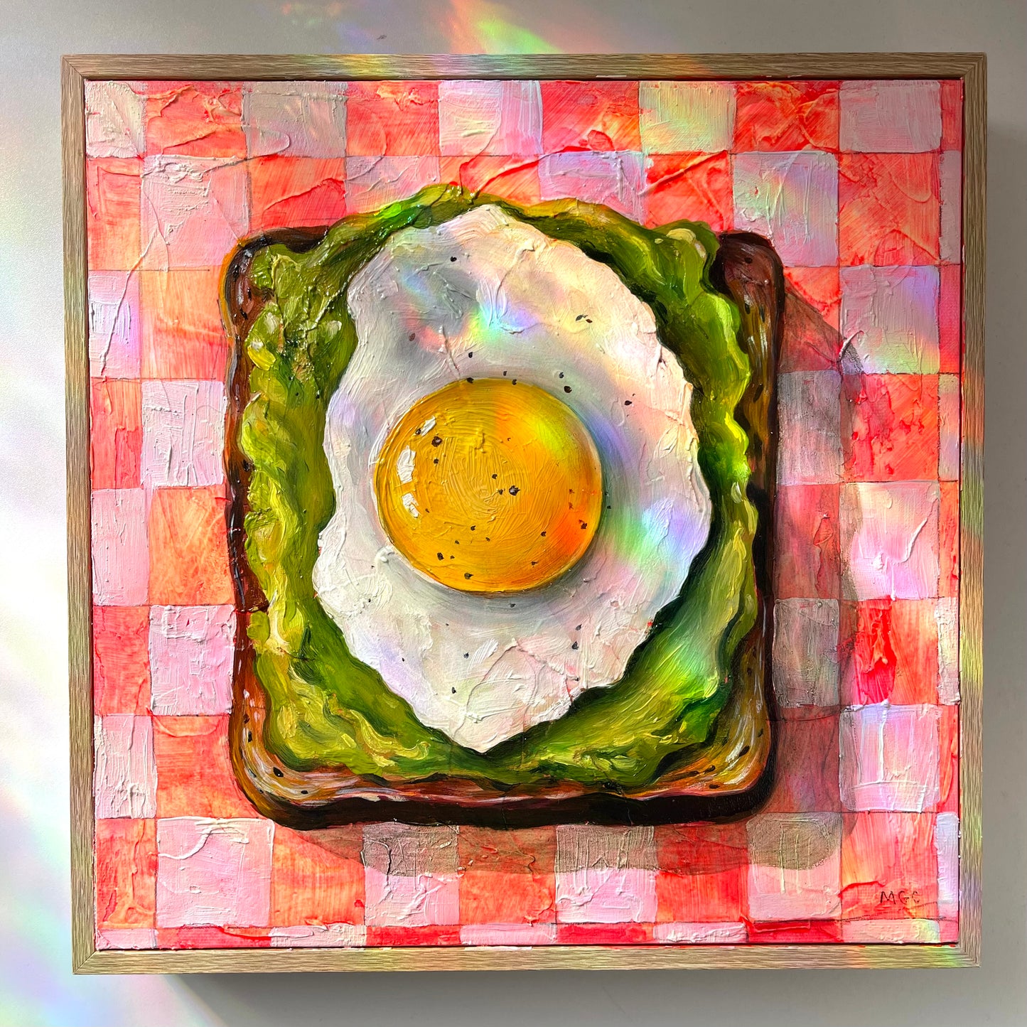 A framed square painting depicting a fried egg on toast over a pink and red checkered pattern, with an iridescent rainbow sheen across the artwork.
