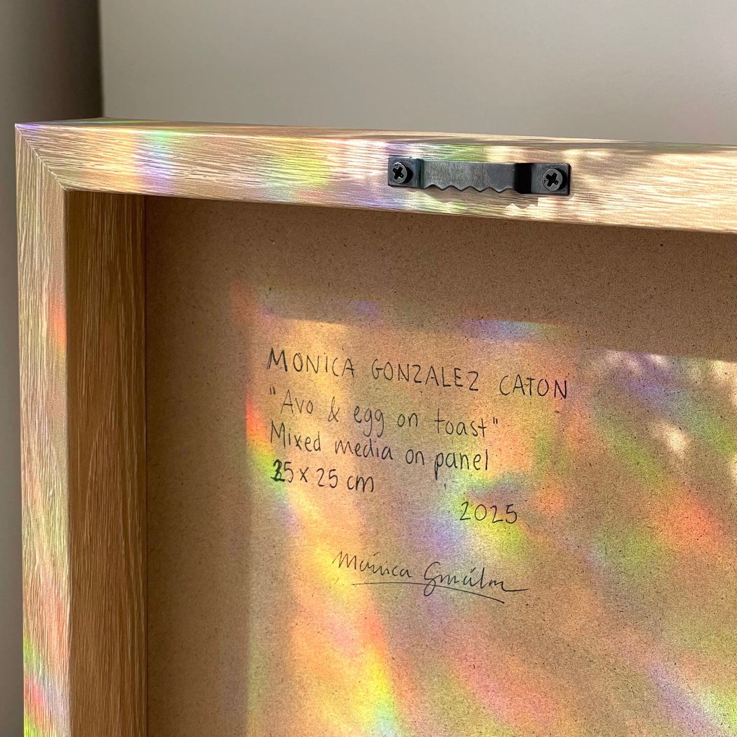 Close-up of the back of a framed artwork with a light wood frame and iridescent reflective surface. Handwritten text on the backing reads 'MONICA GONZALEZ CATON', 'Avo & egg on toast', 'Mixed media on panel', '25 x 25 cm', and '2025', with a signature below.