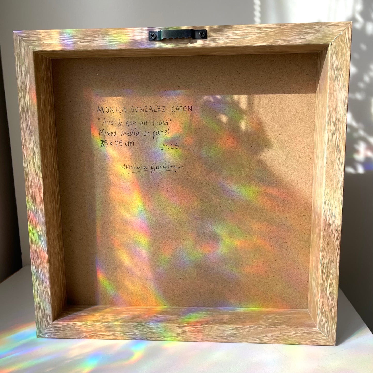 A square wooden shadow box display frame with a cork backing, photographed indoors on a white surface. Sunlight and colored light reflections are cast across the frame and backing.