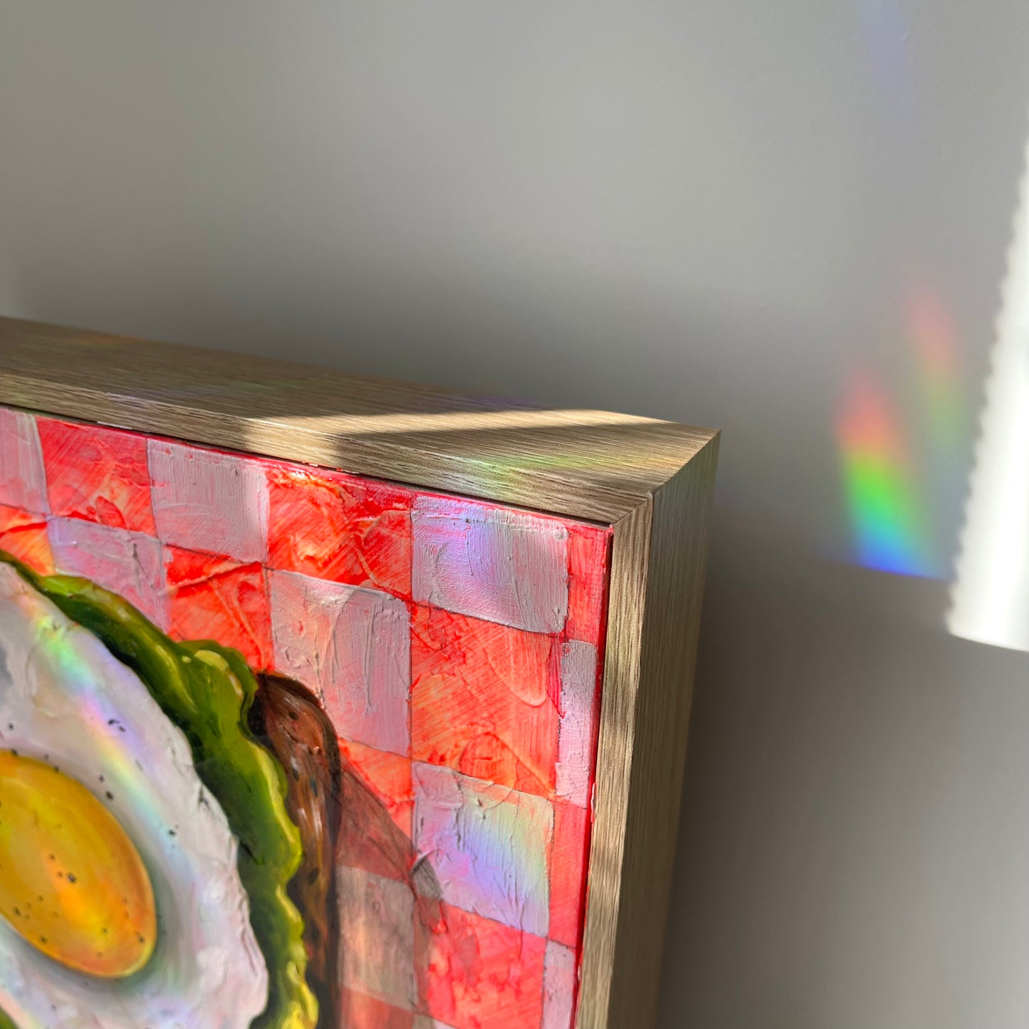 A close-up of a framed artwork showing a textured red and white checkered pattern with a painted fried egg and green vegetable, photographed against a neutral gray wall with sunlight casting a rainbow reflection.