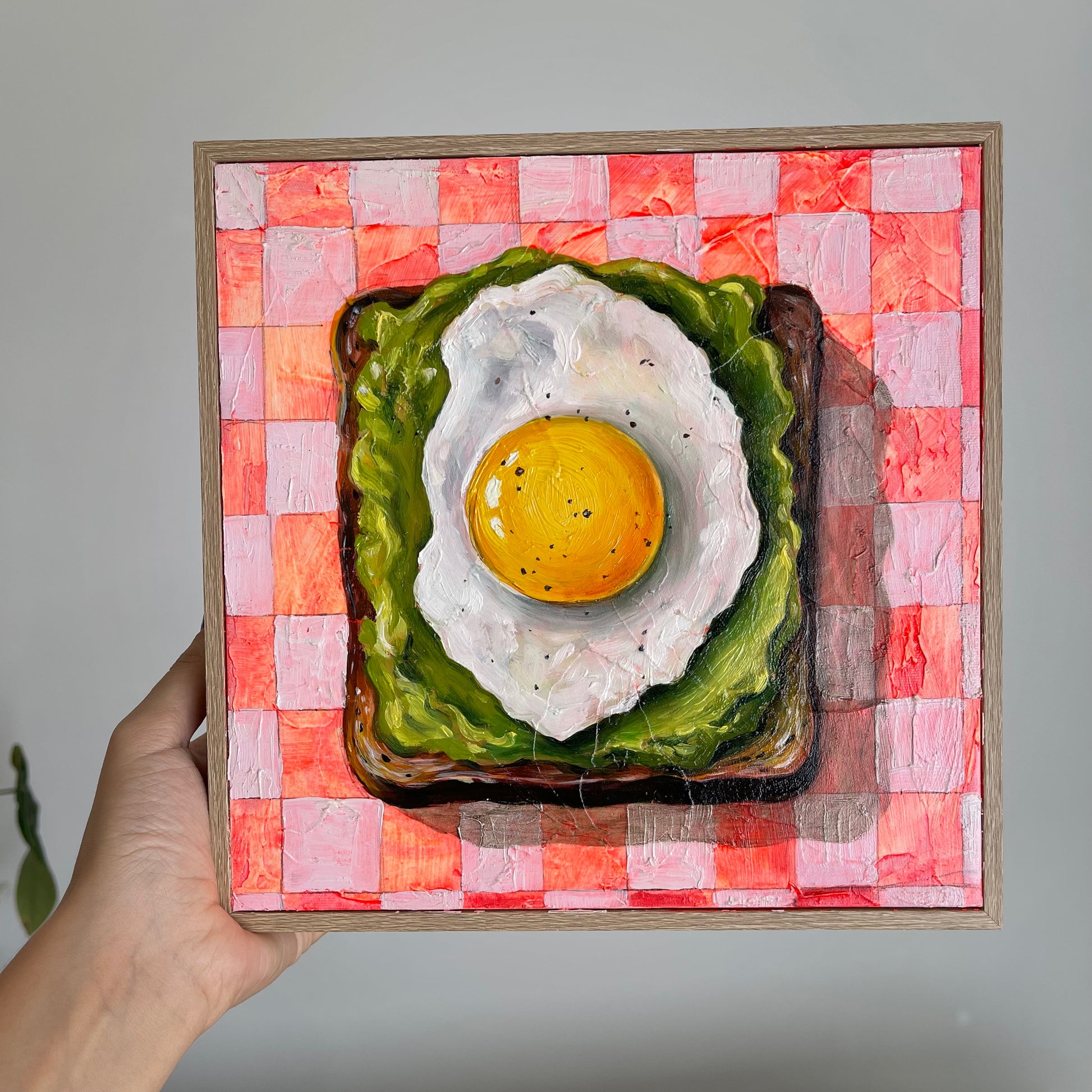 A hand holds a square framed painting depicting a fried egg on toast over a pink and white checkered background. The painting is shown against a plain light gray wall.