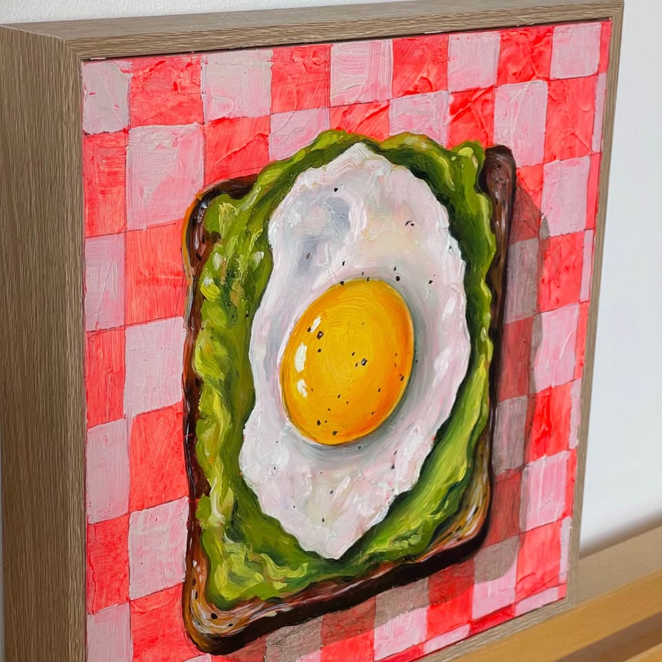 A framed painting of a fried egg on toast against a red and white checkered background, shown in a light wood frame.