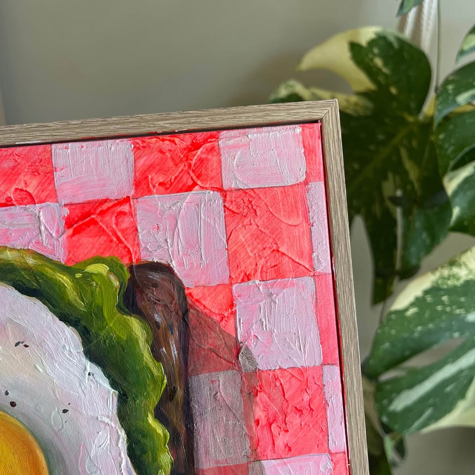 A close-up of a framed painting showing a red and white checkered pattern with green and brown brushstrokes, partially cropped with a variegated houseplant in the background.