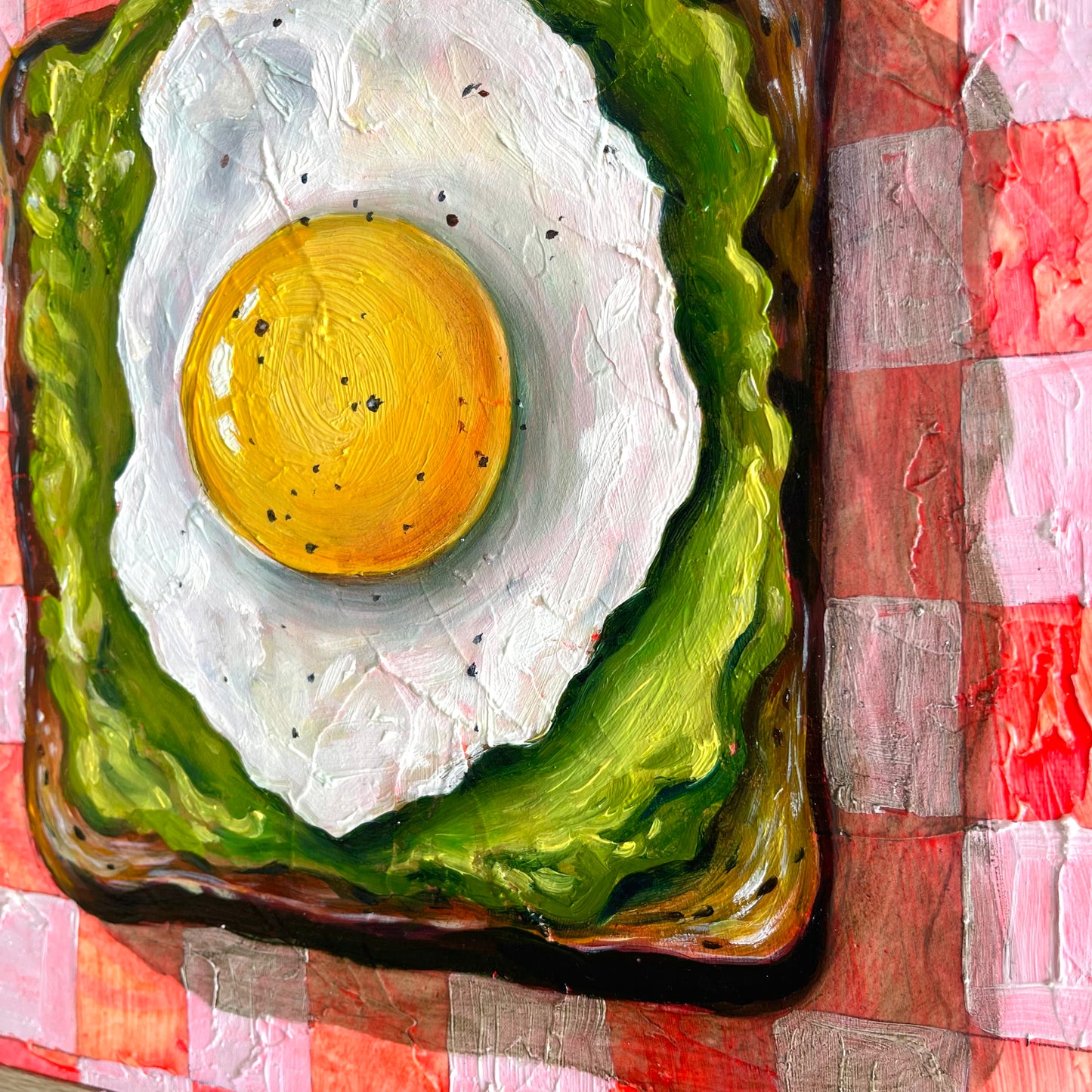 A colorful painting of a fried egg on a slice of toast with green spread, set on a red and white checkered tablecloth background.