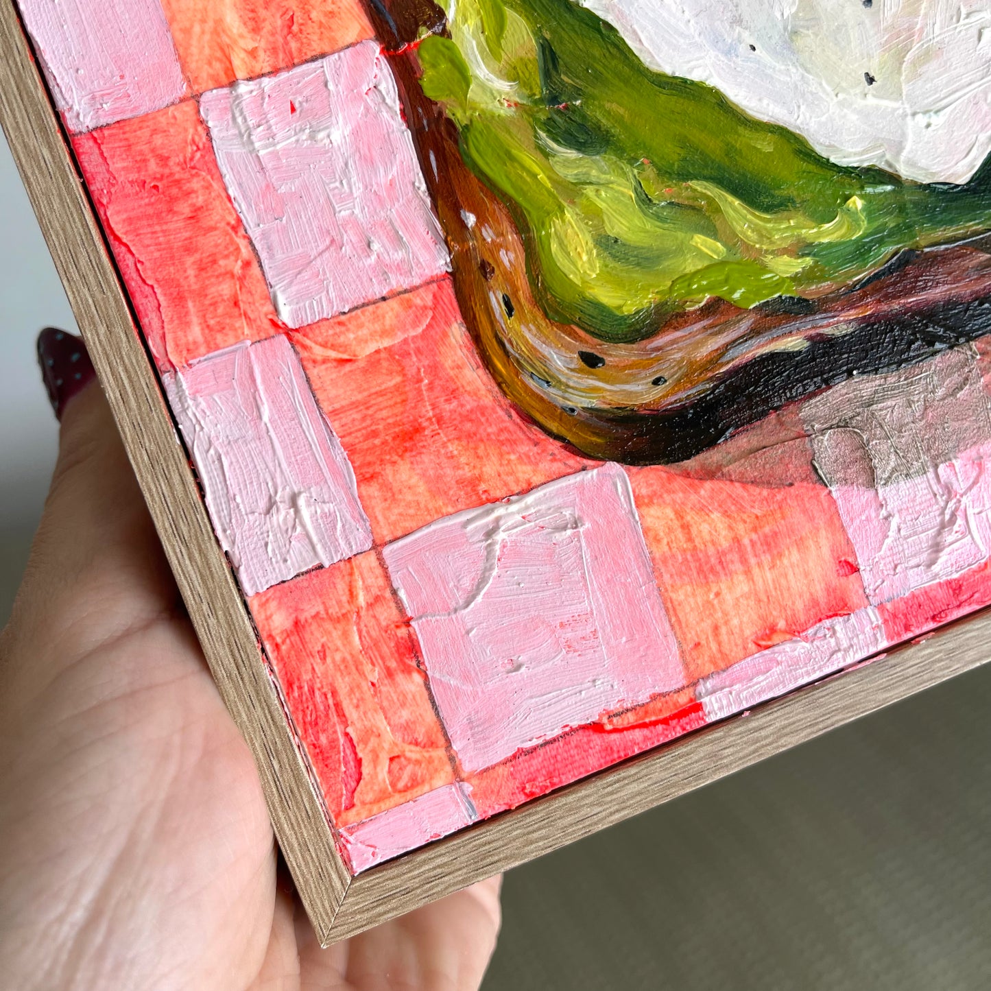 A close-up of a hand holding a framed painting, showing the textured impasto brushstrokes of the artwork. The painting features a patchwork of pink, coral, and red squares, with a green and white textured section in the upper right.
