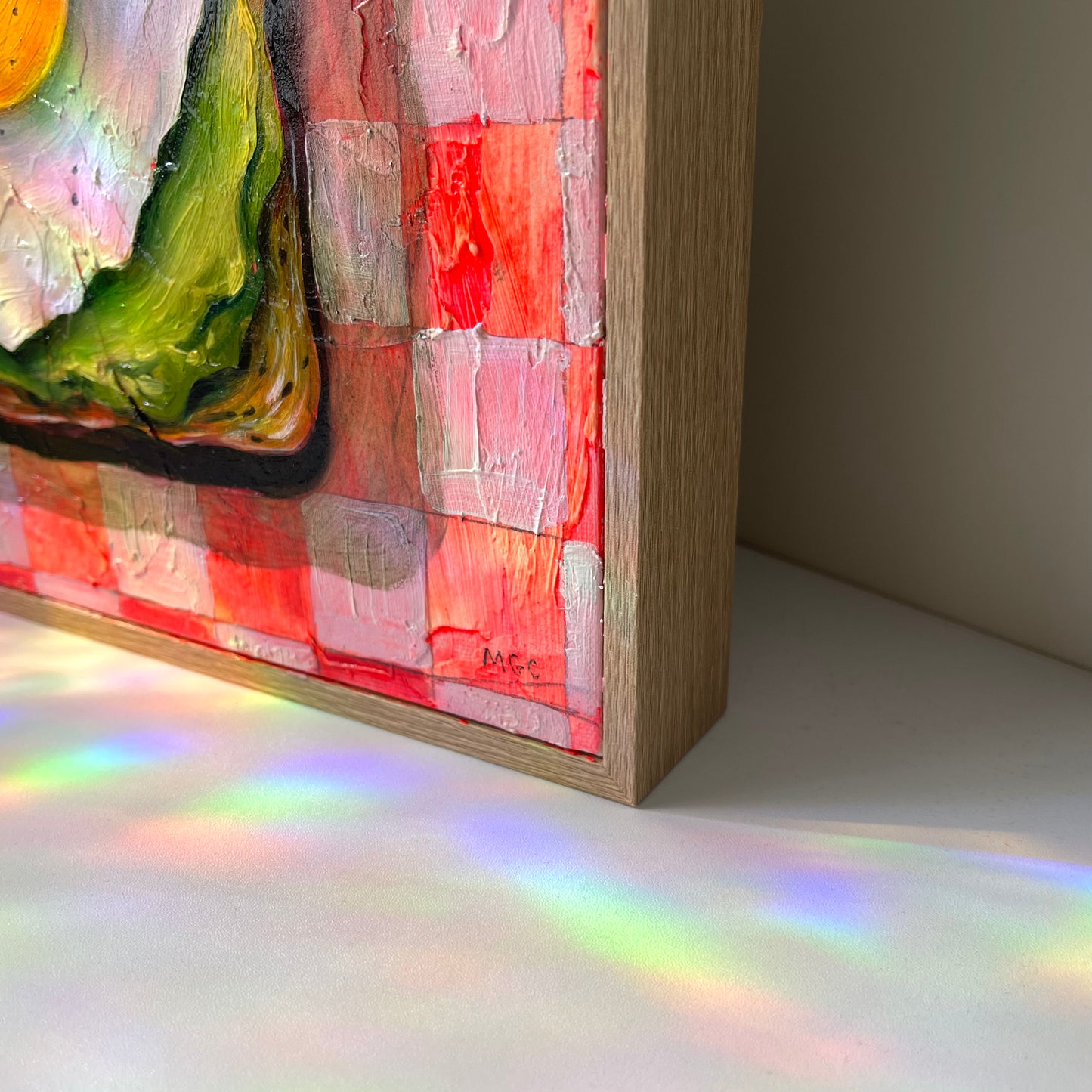 A close-up of a framed textured acrylic painting showing vibrant red, pink, green, and white brushstrokes, casting a rainbow of light onto a white surface below.