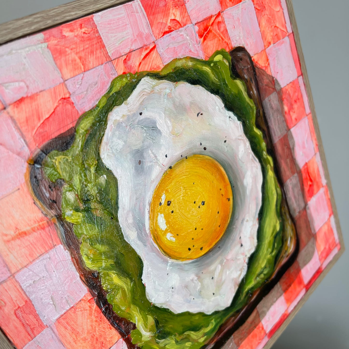 A close-up of a painted wooden box or plaque featuring a fried egg on toast, with a red-and-white checkered surface around it.