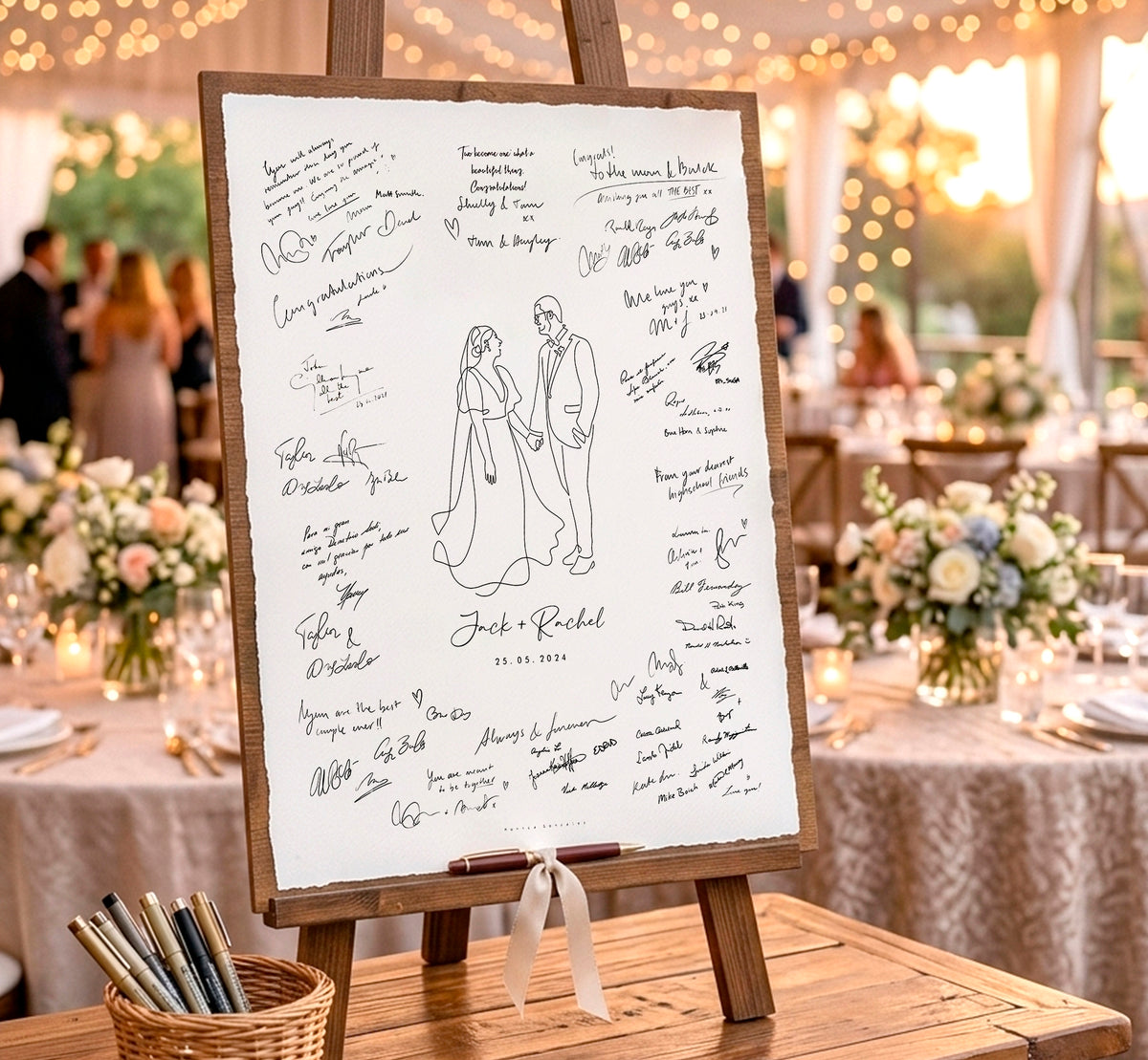A wooden wedding reception sign-on-a-stand with a white board covered in guest signatures and a simple bride-and-groom illustration, set on a table in a warmly lit event venue.