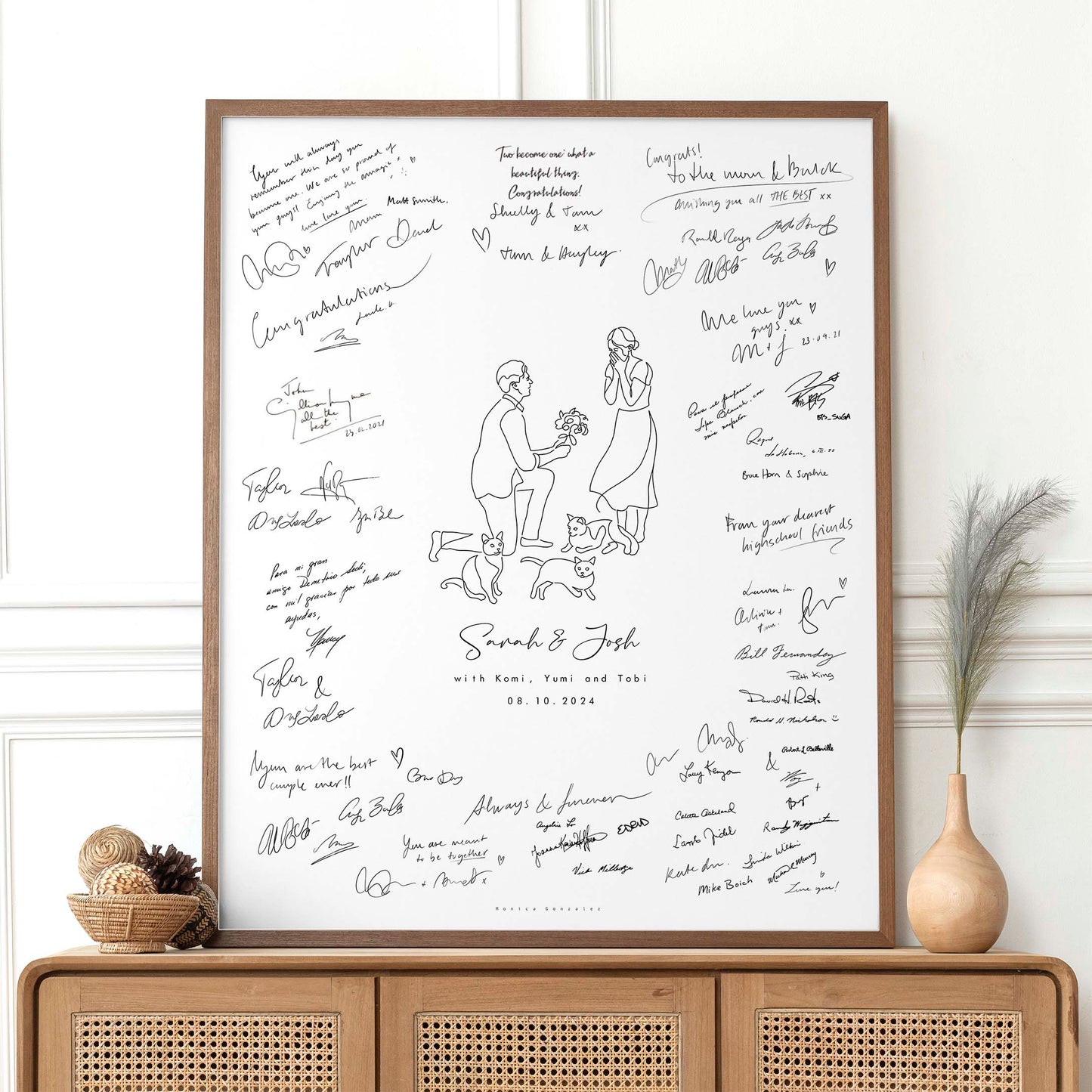 Framed artwork with signatures and a couple illustration on their engagement day along with their three cats on a white wall.
