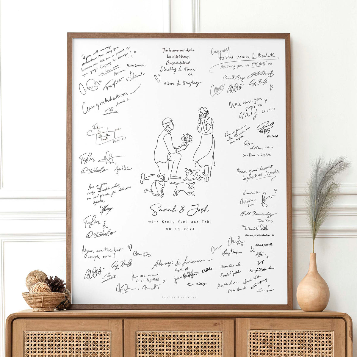 Framed artwork with signatures and a couple illustration on their engagement day along with their three cats on a white wall.