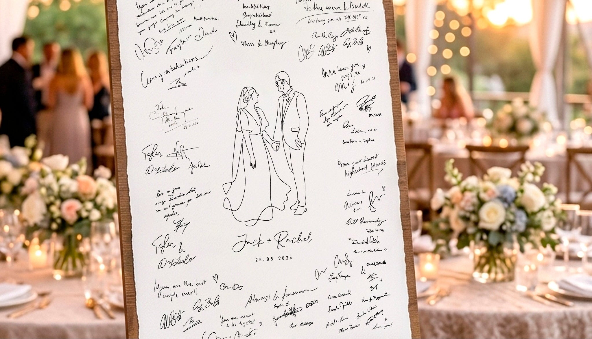 A wooden wedding reception sign-on-a-stand with a white board covered in guest signatures and a simple bride-and-groom illustration, set on a table in a warmly lit event venue.