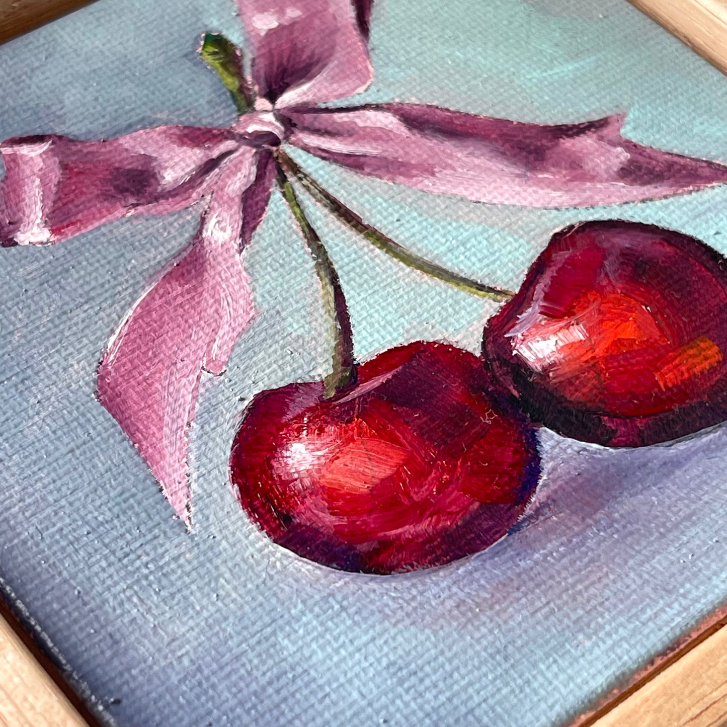 A close-up of a painted artwork showing two red cherries with stems tied by a pink ribbon on a pale blue background.
