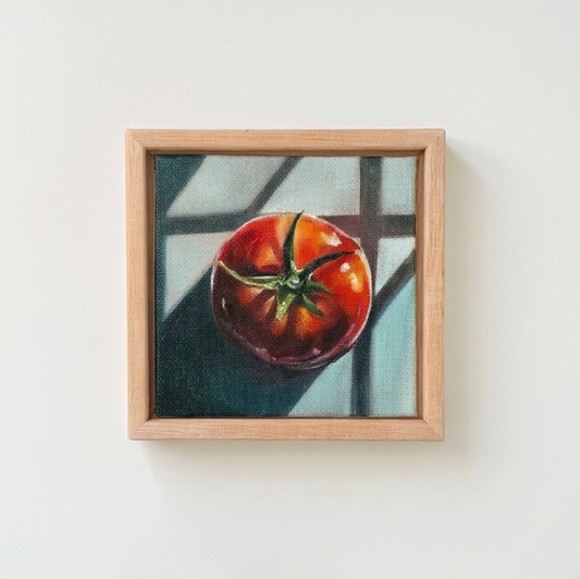 A framed still life painting of a red tomato on a light wood frame, mounted on a plain white wall.