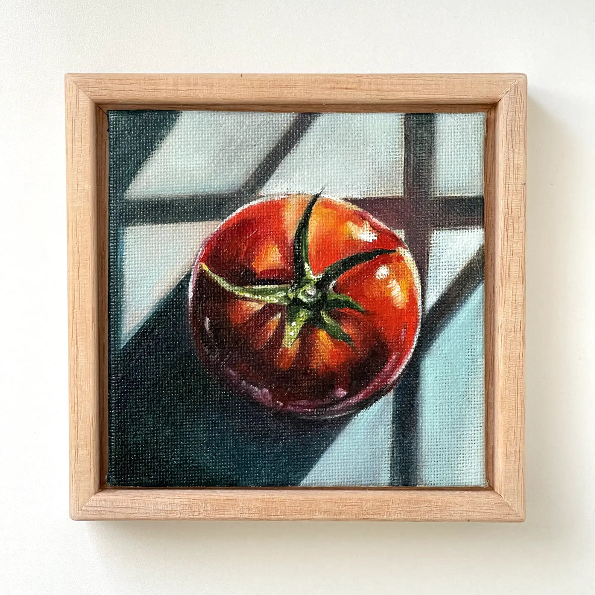 A square framed painting of a red tomato with green stem, set against a dark and light window-pane background, mounted on a plain white wall.