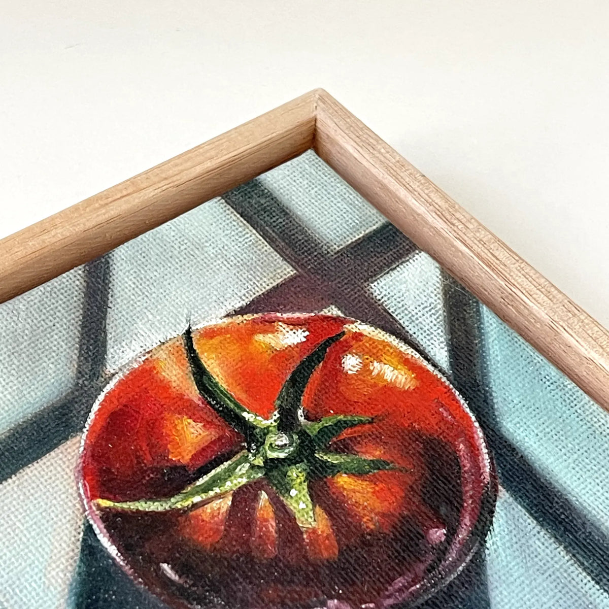 A close-up of a framed painting featuring a red tomato on a blue and gray patterned background, with a light wood frame visible.