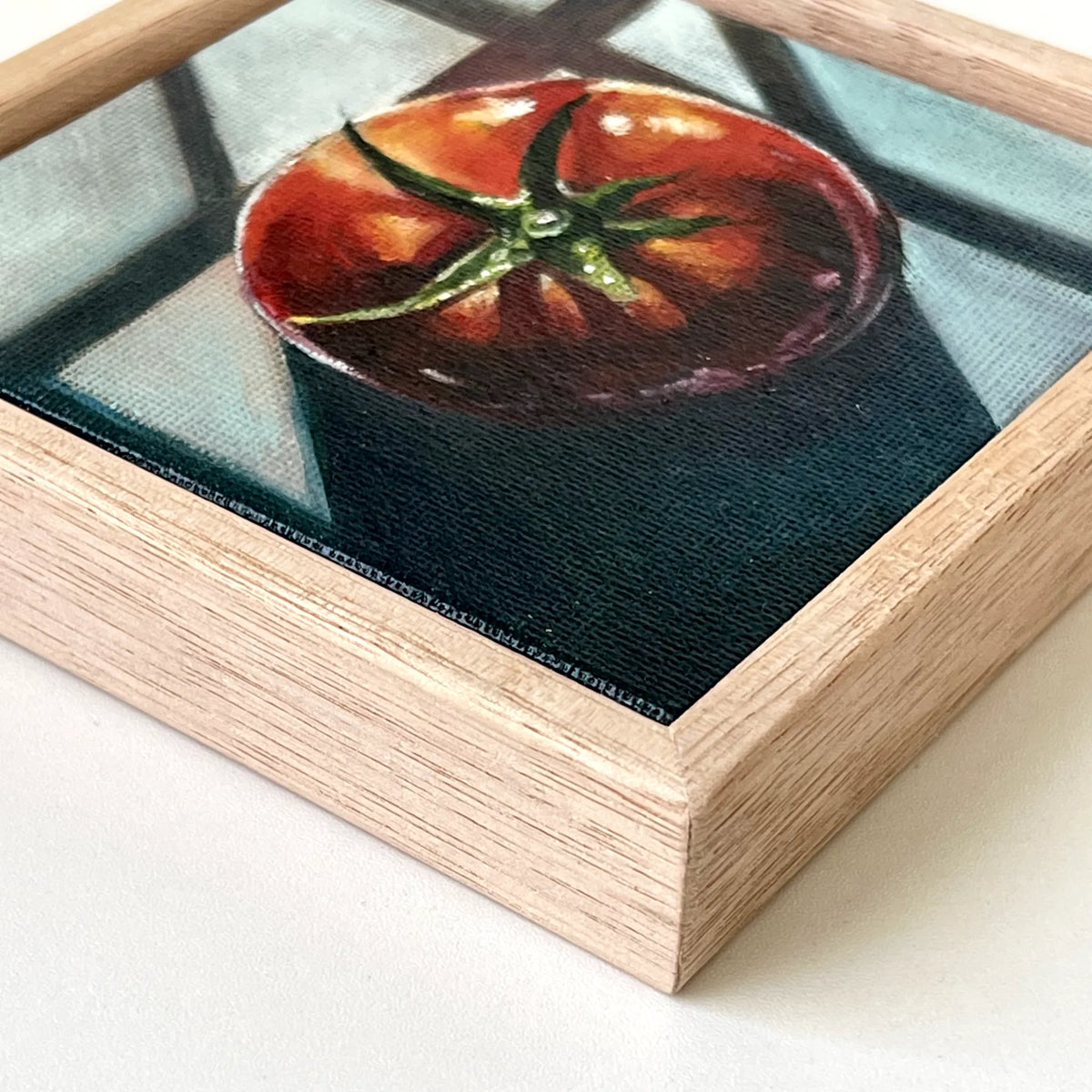 A framed square painting of a red tomato on a dark background, set in a light natural wood frame with a glass or acrylic top surface.