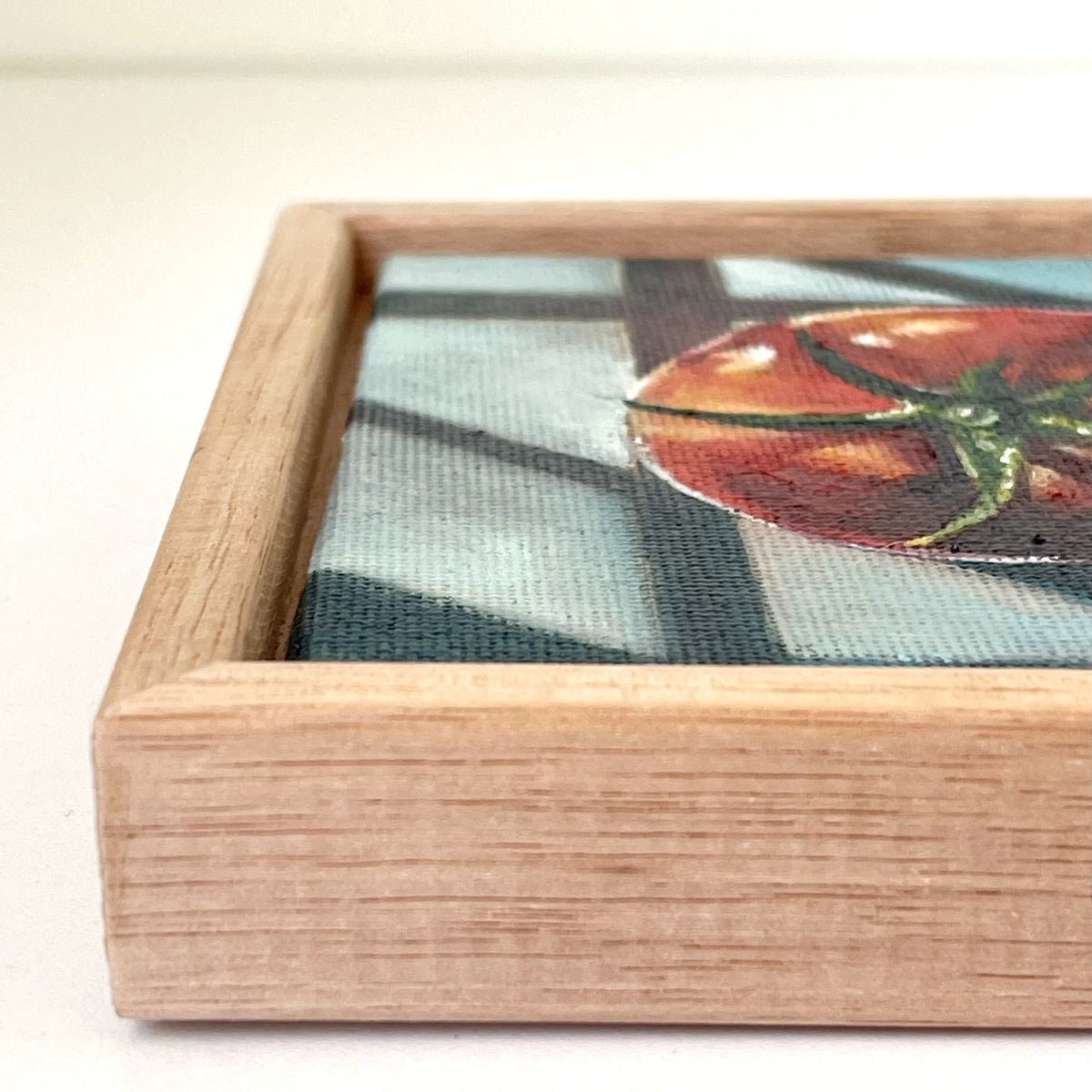 A close-up of a framed artwork showing a painted tomato on a textured background, with a light natural tasmanian oak wood frame visible against a plain light background.