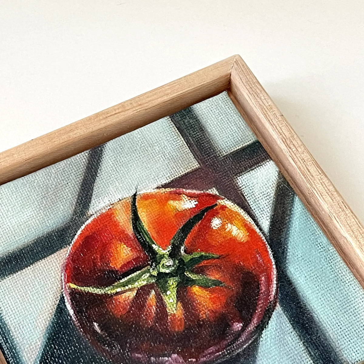 A close-up of a framed painting featuring a red tomato on a blue and gray patterned background, with a light tasmanian oak wood frame visible.