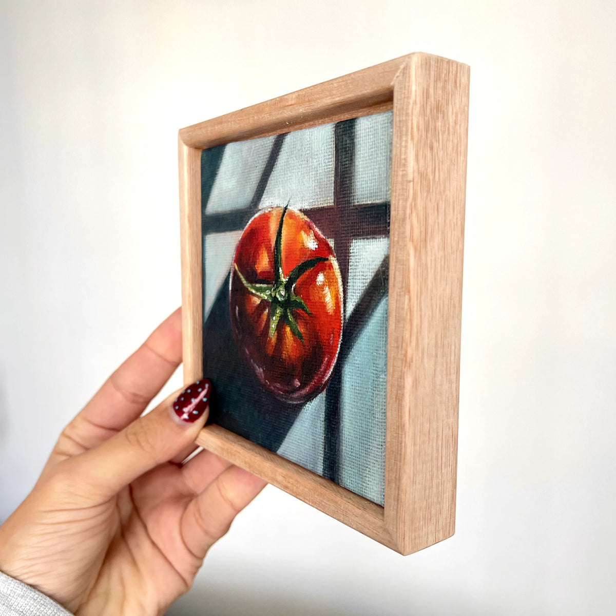 A hand holding a small framed artwork featuring a painted red tomato on a gray background, with a light wood frame.